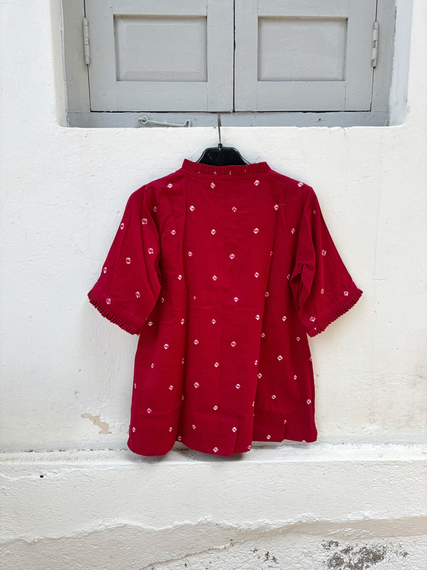 Red cotton satin Bandhani shirt with hand tie-dyed white motifs, pleated round neckline, pleated sleeve hems, coconut shell button closure, relaxed fit, and short side slits. Features Bandhani detailing on both the front and back.