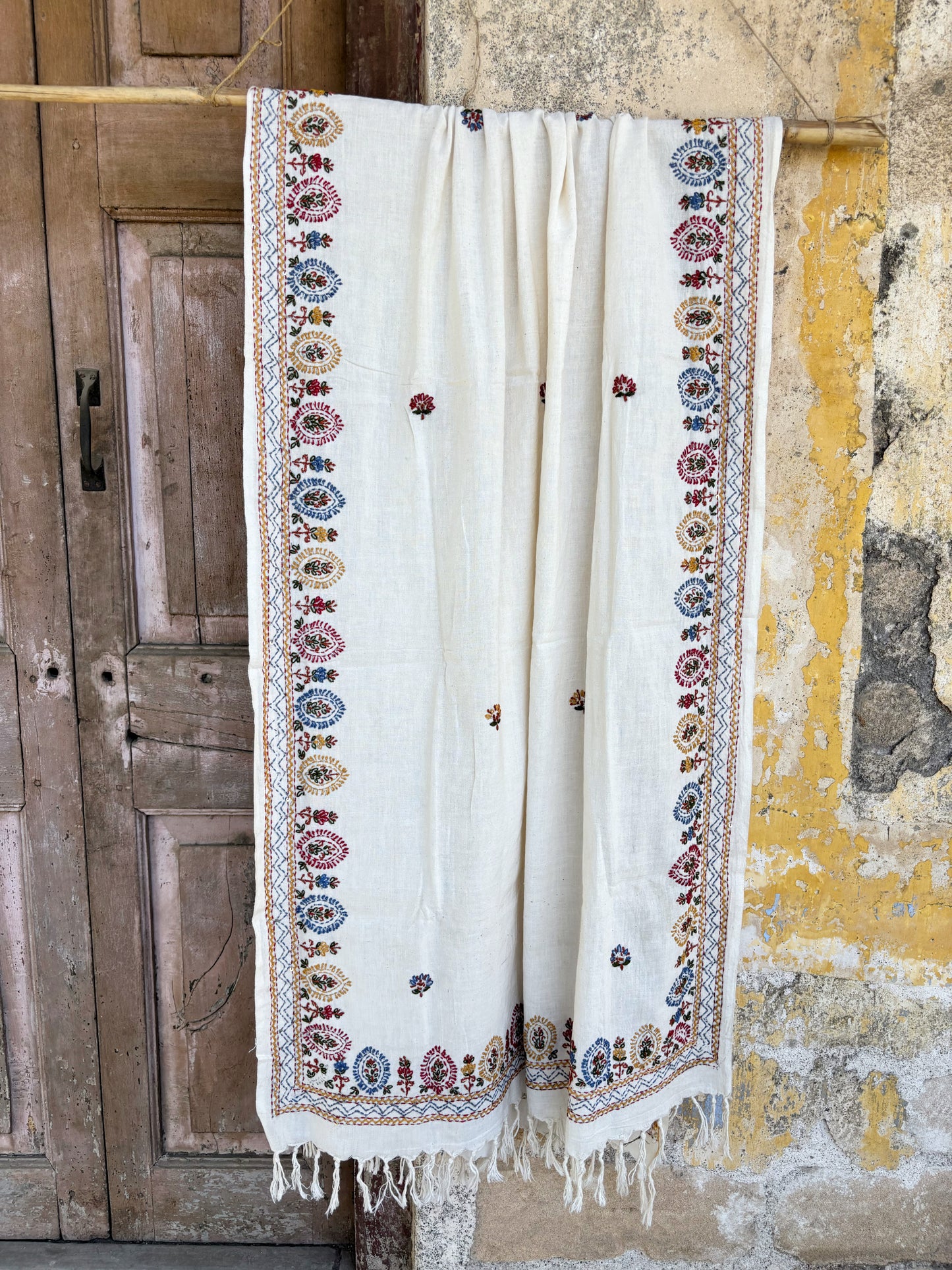 Handspun cotton dupatta in warm ivory shade featuring repeated floral hand embroidered motifs in red, blue, mustard, and green. Lightweight handcrafted dupatta with tassel edges, named Kalika.