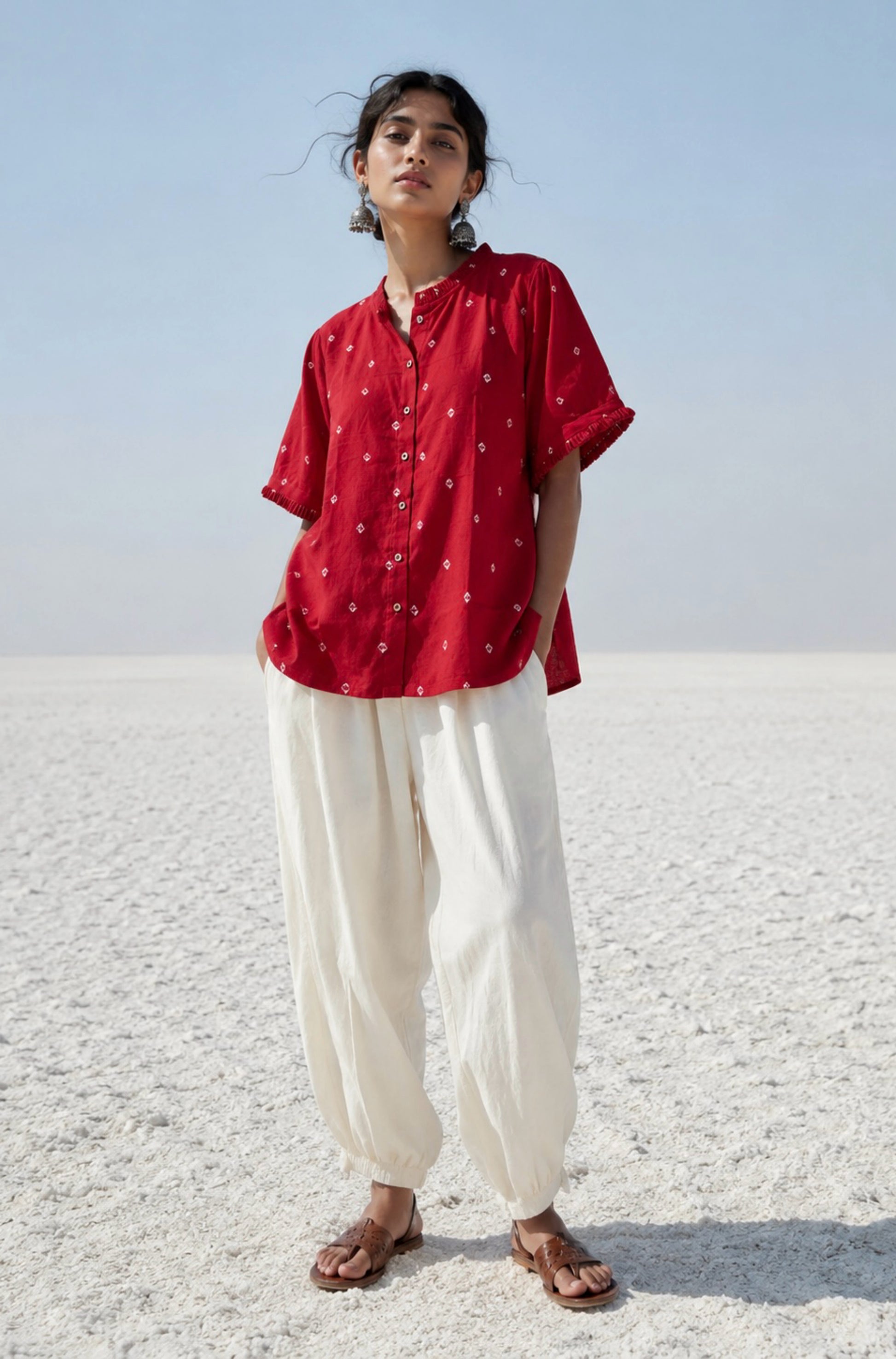 Red cotton satin Bandhani shirt with hand tie-dyed white motifs, pleated round neckline, pleated sleeve hems, coconut shell button closure, relaxed fit, and short side slits. Features Bandhani detailing on both the front and back.