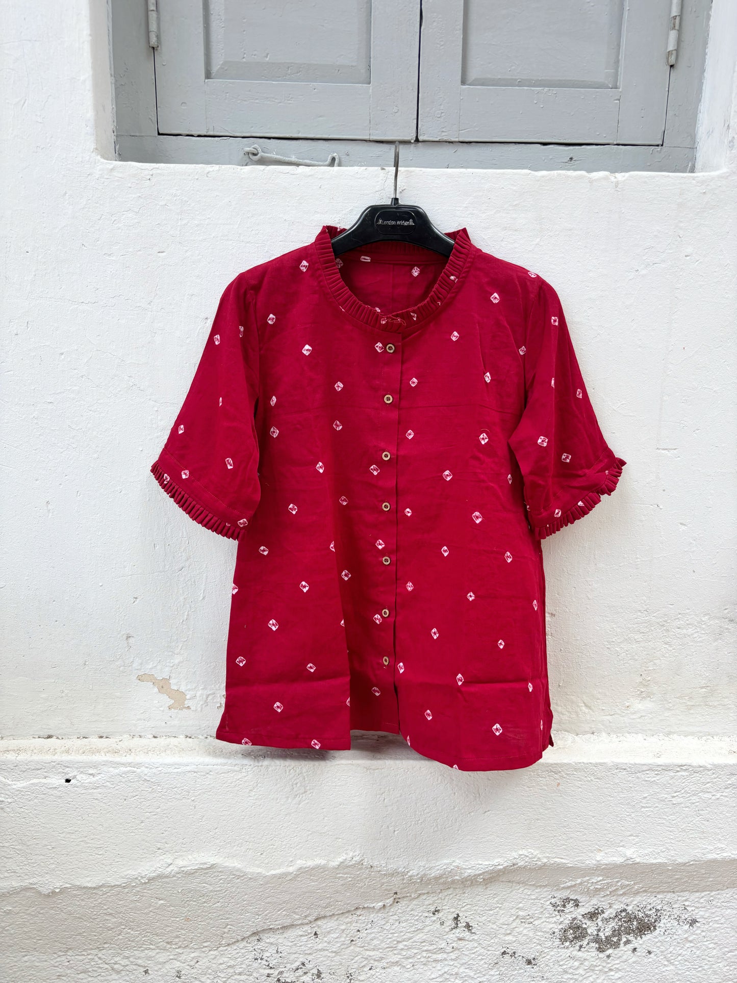 Red cotton satin Bandhani shirt with hand tie-dyed white motifs, pleated round neckline, pleated sleeve hems, coconut shell button closure, relaxed fit, and short side slits. Features Bandhani detailing on both the front and back.