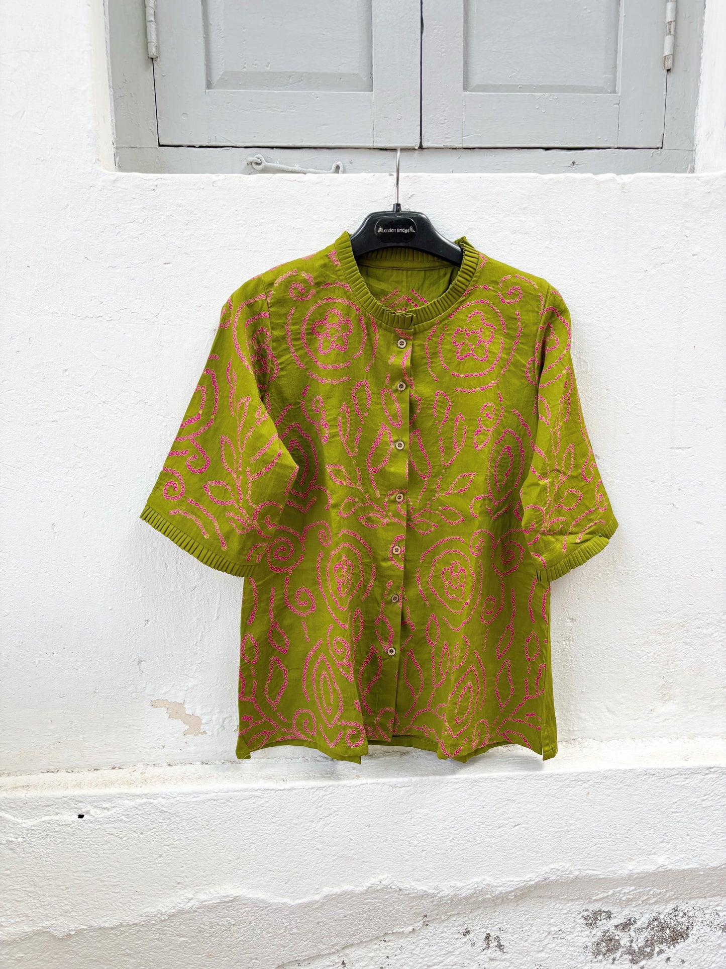 Olive green Jhankar Bandhani cotton shirt with rani pink floral and paisley motifs, pleated neckline, pleated sleeve hems, coconut shell buttons, and relaxed fit.