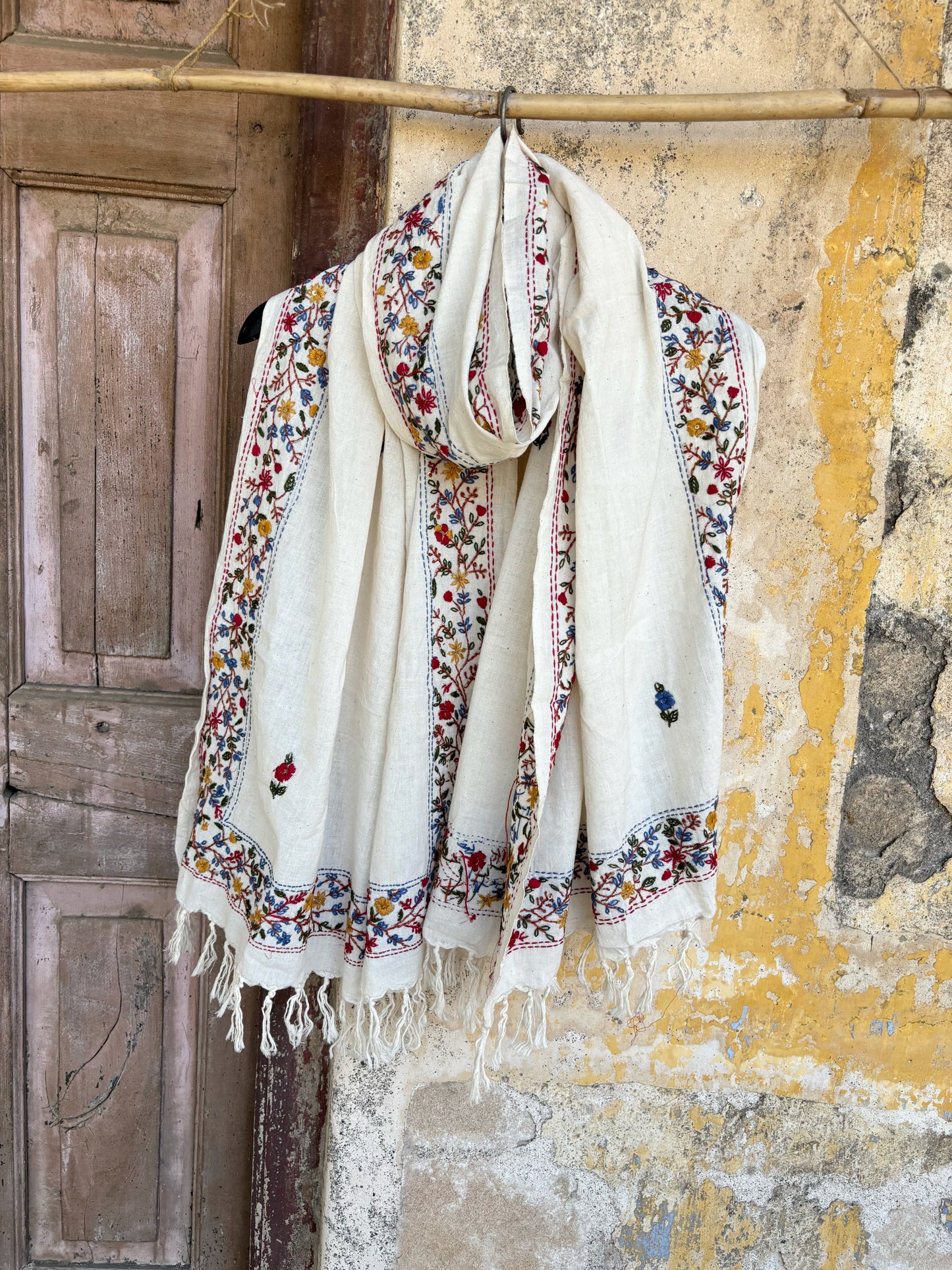 Handspun cotton dupatta in warm ivory shade featuring continuous floral hand embroidery along the borders in red, blue, mustard, and green. Lightweight handcrafted dupatta with tassel edges, named Pallavi.